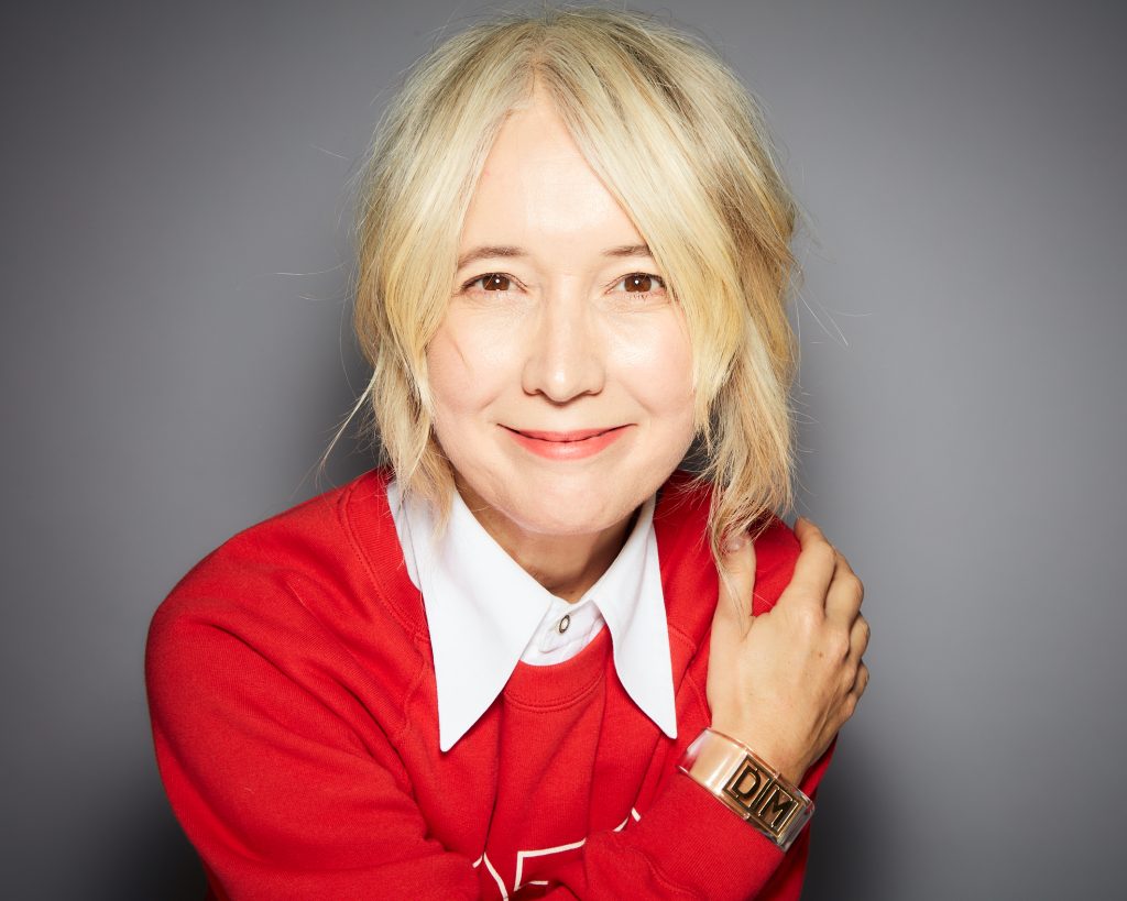 An image of a woman, Justine Simons, smiling. She is wearing a red jumper and white collared shirt.