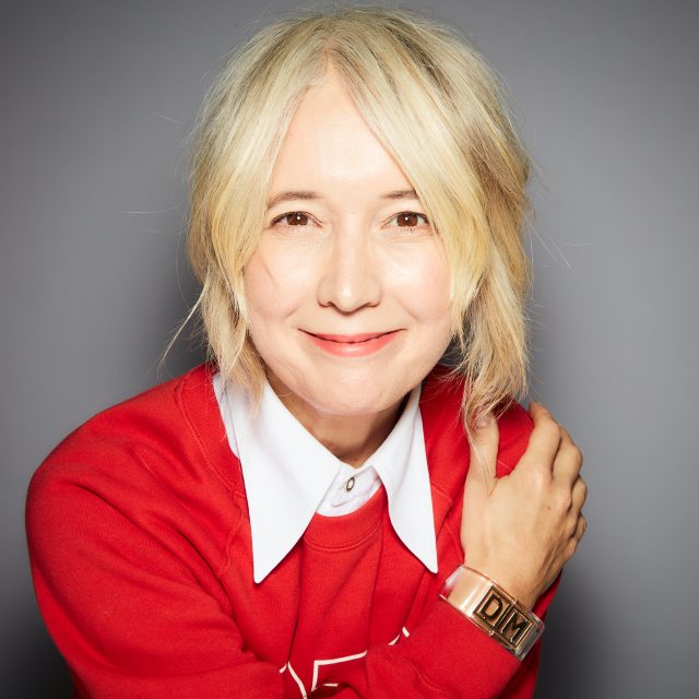 An image of a woman, Justine Simons, smiling. She is wearing a red jumper and white collared shirt.
