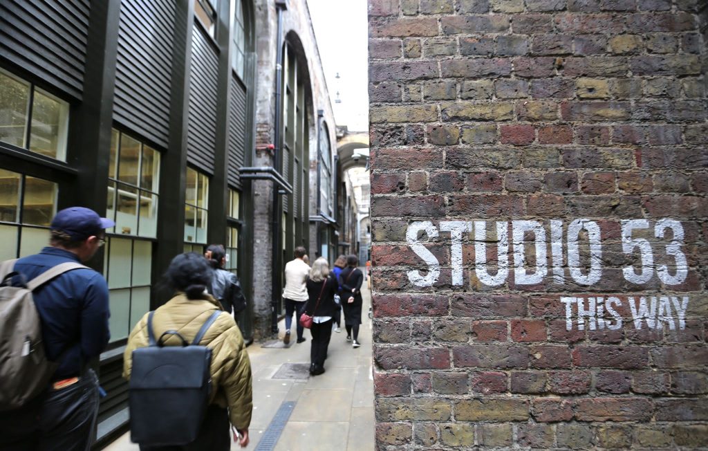 A brick wall reads 'studio 53 this way,' with people walking passed, next to a building.
